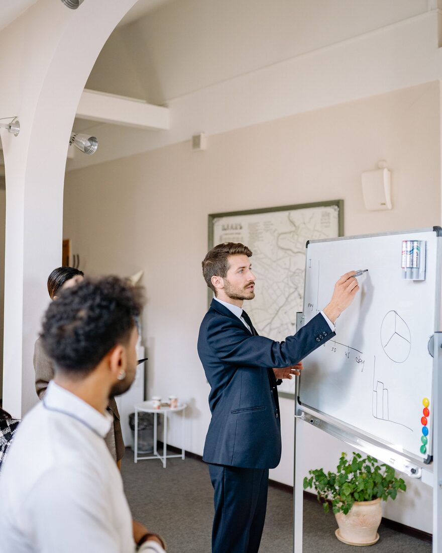 Berater erläutert Kennzahlen an einem Whiteboard vor zwei Zuhörenden